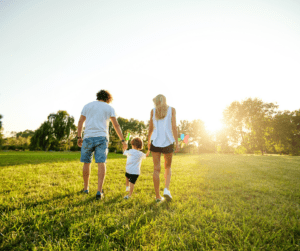 Parents hold the hands of a toddler walking in a field.