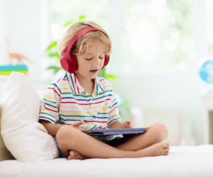 A young blond boy with read headphones plays on a tablet.